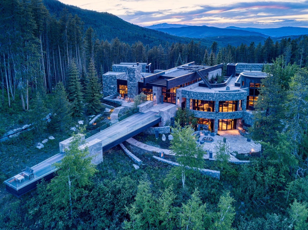 Stone estate at dusk in a forested mountain valley, interior lights just coming on